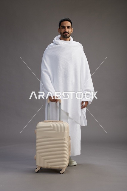 رجل  عربي سعودي خليجي بملابس الإحرام مع حقيبة سفر, الذهاب الى مكه لاداء مناسك الحج والعمره, الجسم بالطول الكامل, خلفيه رماديه