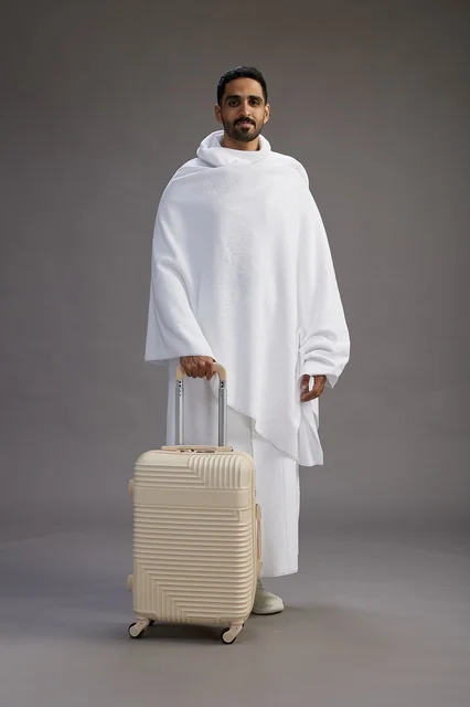 A Saudi Arabian Gulf man in Ihram clothing with a travel bag, going to Mecca to perform the rituals of Hajj and Umrah, full-body, gray background.