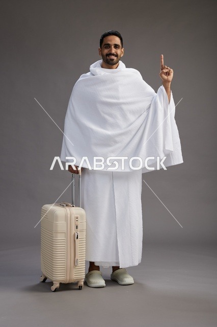 رجل  عربي سعودي خليجي بملابس الإحرام مع حقيبة سفر, الذهاب الى مكه لاداء مناسك الحج والعمره, الجسم بالطول الكامل, خلفيه رماديه