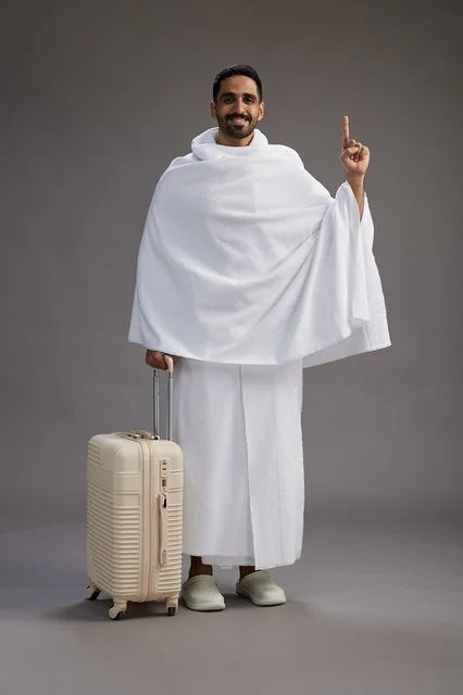 A Saudi Arabian Gulf man in Ihram clothing with a travel bag, going to Mecca to perform the rituals of Hajj and Umrah, full-body, gray background.