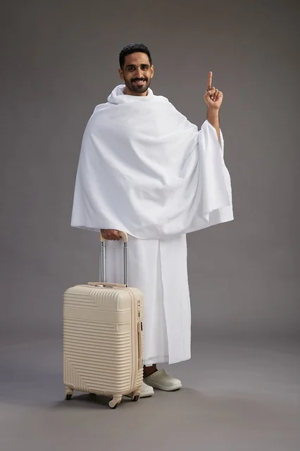A Saudi Arabian Gulf man in Ihram clothing with a travel bag, going to Mecca to perform the rituals of Hajj and Umrah, full-body, gray background.