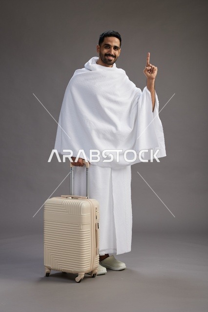 رجل  عربي سعودي خليجي بملابس الإحرام مع حقيبة سفر, الذهاب الى مكه لاداء مناسك الحج والعمره, الجسم بالطول الكامل, خلفيه رماديه