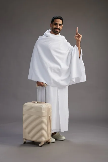 رجل  عربي سعودي خليجي بملابس الإحرام مع حقيبة سفر, الذهاب الى مكه لاداء مناسك الحج والعمره, الجسم بالطول الكامل, خلفيه رماديه