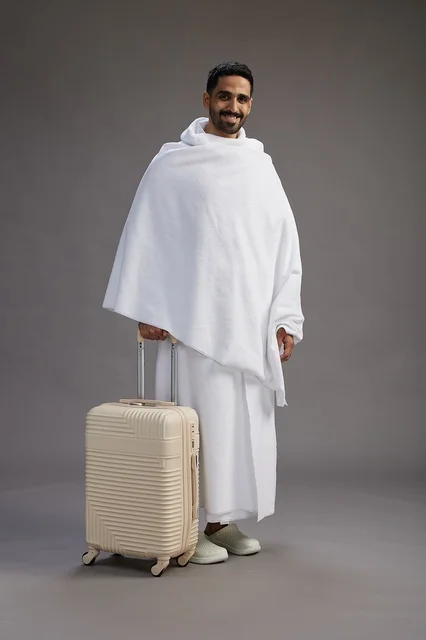 A Saudi Arabian Gulf man in Ihram clothing with a travel bag, going to Mecca to perform the rituals of Hajj and Umrah, full-body, gray background.