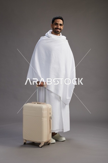 رجل  عربي سعودي خليجي بملابس الإحرام مع حقيبة سفر, الذهاب الى مكه لاداء مناسك الحج والعمره, الجسم بالطول الكامل, خلفيه رماديه