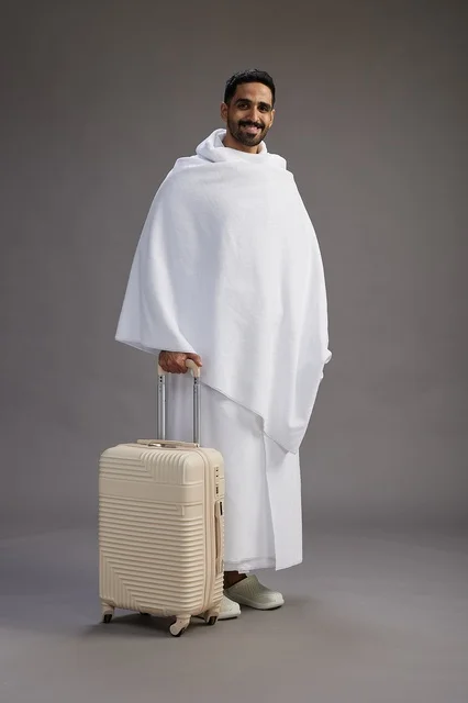 A Saudi Arabian Gulf man in Ihram clothing with a travel bag, going to Mecca to perform the rituals of Hajj and Umrah, full-body, gray background.