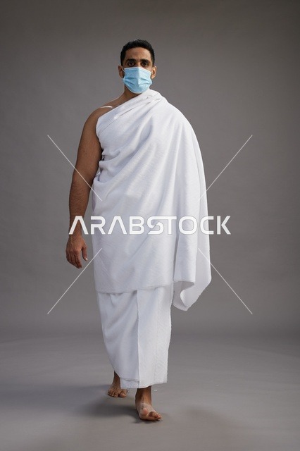 رجل عربي  سعودي  خليجي بملابس الإحرام يرتدي كمامة واقف استعدادًا للحج, الوقايه من فايروس كورونا, كامل الجسم, خلفيه رماديه