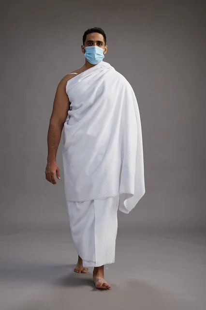 رجل عربي  سعودي  خليجي بملابس الإحرام يرتدي كمامة واقف استعدادًا للحج, الوقايه من فايروس كورونا, كامل الجسم, خلفيه رماديه