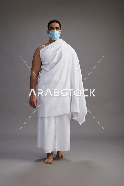 رجل عربي  سعودي  خليجي بملابس الإحرام يرتدي كمامة واقف استعدادًا للحج, الوقايه من فايروس كورونا, كامل الجسم, خلفيه رماديه