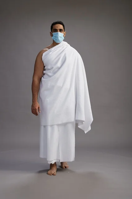 رجل عربي  سعودي  خليجي بملابس الإحرام يرتدي كمامة واقف استعدادًا للحج, الوقايه من فايروس كورونا, كامل الجسم, خلفيه رماديه