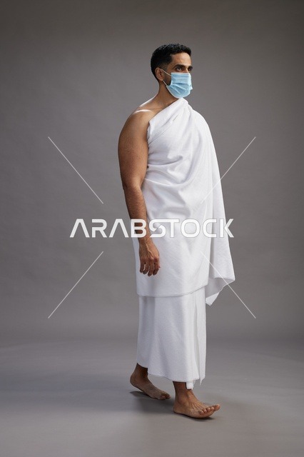 رجل عربي  سعودي  خليجي بملابس الإحرام يرتدي كمامة واقف استعدادًا للحج, الوقايه من فايروس كورونا, كامل الجسم, خلفيه رماديه