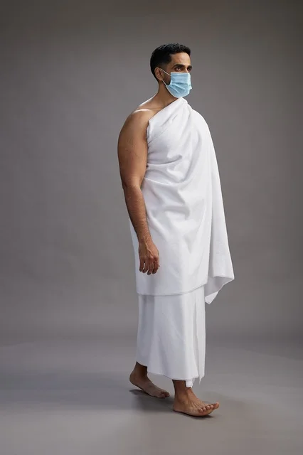 An Arab Saudi Gulf man in Ihram clothing wearing a mask standing ready for Hajj, protection from the coronavirus, full body, gray background.