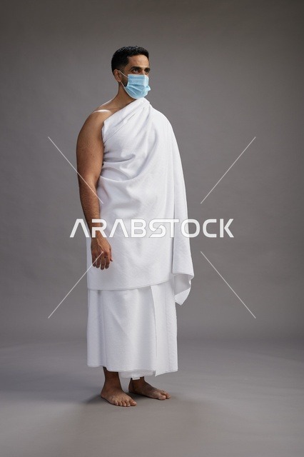 رجل عربي  سعودي  خليجي بملابس الإحرام يرتدي كمامة واقف استعدادًا للحج, الوقايه من فايروس كورونا, كامل الجسم, خلفيه رماديه