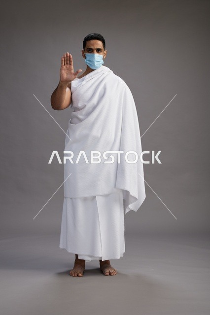 رجل عربي  سعودي  خليجي بملابس الإحرام يرتدي كمامة واقف استعدادًا للحج, الوقايه من فايروس كورونا, كامل الجسم, خلفيه رماديه