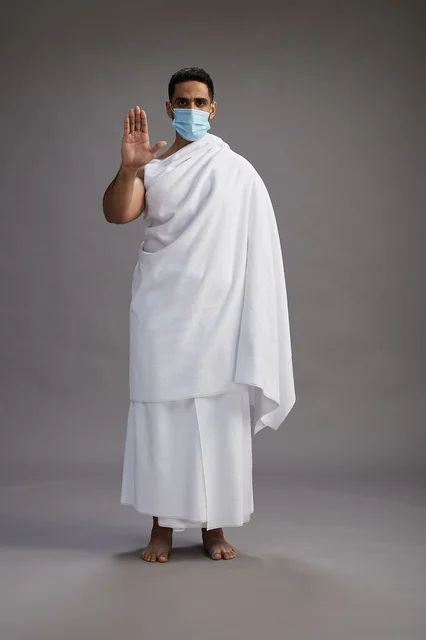 An Arab Saudi Gulf man in Ihram clothing wearing a mask standing ready for Hajj, protection from the coronavirus, full body, gray background.