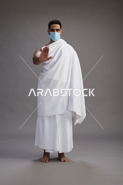 رجل عربي  سعودي  خليجي بملابس الإحرام يرتدي كمامة واقف استعدادًا للحج, الوقايه من فايروس كورونا, كامل الجسم, خلفيه رماديه