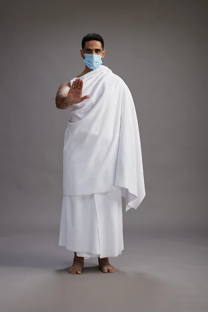 رجل عربي  سعودي  خليجي بملابس الإحرام يرتدي كمامة واقف استعدادًا للحج, الوقايه من فايروس كورونا, كامل الجسم, خلفيه رماديه