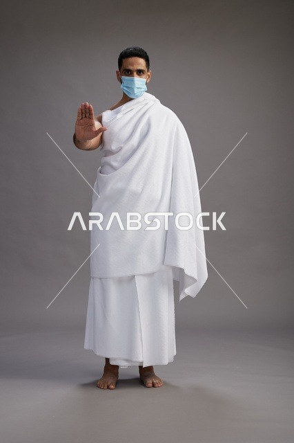 رجل عربي  سعودي  خليجي بملابس الإحرام يرتدي كمامة واقف استعدادًا للحج, الوقايه من فايروس كورونا, كامل الجسم, خلفيه رماديه