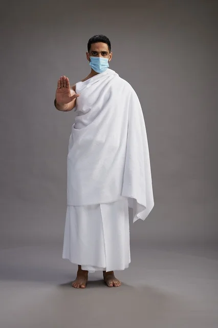 رجل عربي  سعودي  خليجي بملابس الإحرام يرتدي كمامة واقف استعدادًا للحج, الوقايه من فايروس كورونا, كامل الجسم, خلفيه رماديه