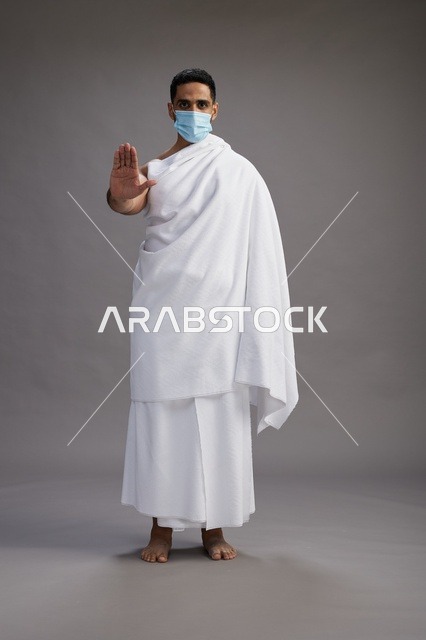 رجل عربي  سعودي  خليجي بملابس الإحرام يرتدي كمامة واقف استعدادًا للحج, الوقايه من فايروس كورونا, كامل الجسم, خلفيه رماديه