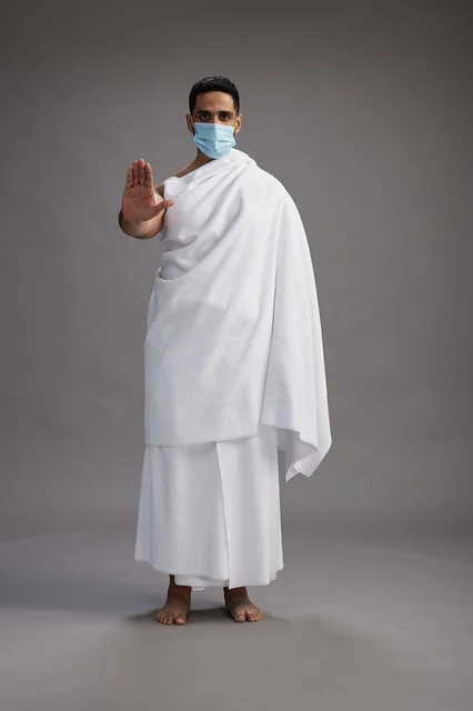 رجل عربي  سعودي  خليجي بملابس الإحرام يرتدي كمامة واقف استعدادًا للحج, الوقايه من فايروس كورونا, كامل الجسم, خلفيه رماديه