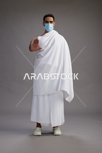 رجل عربي  سعودي  خليجي بملابس الإحرام يرتدي كمامة واقف استعدادًا للحج, الوقايه من فايروس كورونا, كامل الجسم, خلفيه رماديه