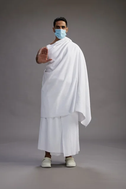 An Arab Saudi Gulf man in Ihram clothing wearing a mask standing ready for Hajj, protection from the coronavirus, full body, gray background.