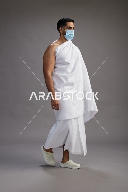 رجل عربي  سعودي  خليجي بملابس الإحرام يرتدي كمامة واقف استعدادًا للحج, الوقايه من فايروس كورونا, كامل الجسم, خلفيه رماديه