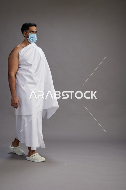 رجل عربي  سعودي  خليجي بملابس الإحرام يرتدي كمامة واقف استعدادًا للحج, الوقايه من فايروس كورونا, كامل الجسم, خلفيه رماديه