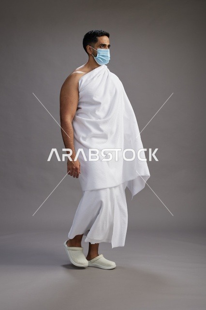 رجل عربي  سعودي  خليجي بملابس الإحرام يرتدي كمامة واقف استعدادًا للحج, الوقايه من فايروس كورونا, كامل الجسم, خلفيه رماديه