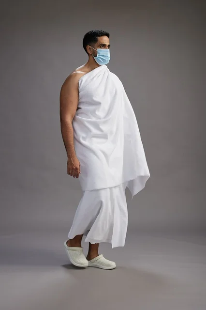 رجل عربي  سعودي  خليجي بملابس الإحرام يرتدي كمامة واقف استعدادًا للحج, الوقايه من فايروس كورونا, كامل الجسم, خلفيه رماديه
