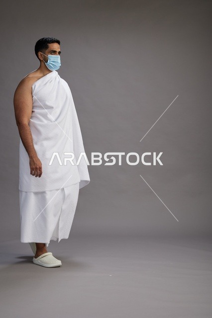 رجل عربي  سعودي  خليجي بملابس الإحرام يرتدي كمامة واقف استعدادًا للحج, الوقايه من فايروس كورونا, كامل الجسم, خلفيه رماديه