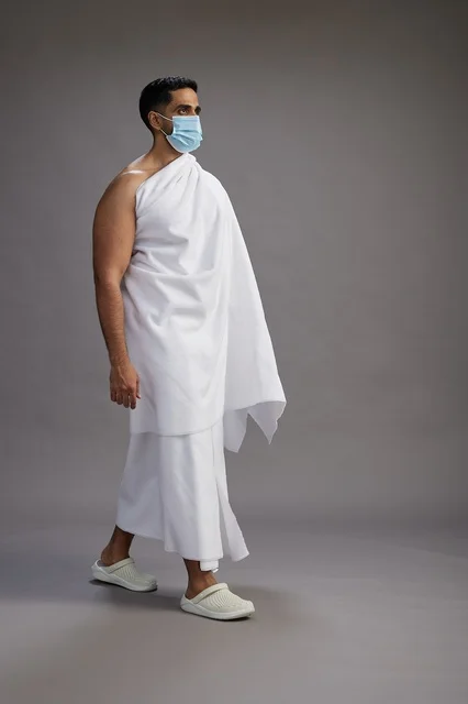 رجل عربي  سعودي  خليجي بملابس الإحرام يرتدي كمامة واقف استعدادًا للحج, الوقايه من فايروس كورونا, كامل الجسم, خلفيه رماديه