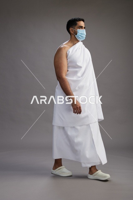 رجل عربي  سعودي  خليجي بملابس الإحرام يرتدي كمامة واقف استعدادًا للحج, الوقايه من فايروس كورونا, كامل الجسم, خلفيه رماديه
