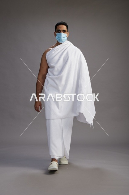 رجل عربي  سعودي  خليجي بملابس الإحرام يرتدي كمامة واقف استعدادًا للحج, الوقايه من فايروس كورونا, كامل الجسم, خلفيه رماديه