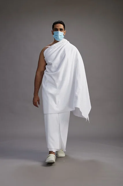 An Arab Saudi Gulf man in Ihram clothing wearing a mask standing ready for Hajj, protection from the coronavirus, full body, gray background.
