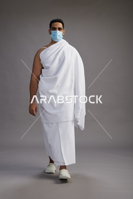 رجل عربي  سعودي  خليجي بملابس الإحرام يرتدي كمامة واقف استعدادًا للحج, الوقايه من فايروس كورونا, كامل الجسم, خلفيه رماديه