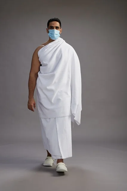 رجل عربي  سعودي  خليجي بملابس الإحرام يرتدي كمامة واقف استعدادًا للحج, الوقايه من فايروس كورونا, كامل الجسم, خلفيه رماديه