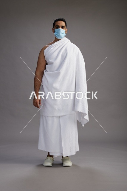 رجل عربي  سعودي  خليجي بملابس الإحرام يرتدي كمامة واقف استعدادًا للحج, الوقايه من فايروس كورونا, كامل الجسم, خلفيه رماديه