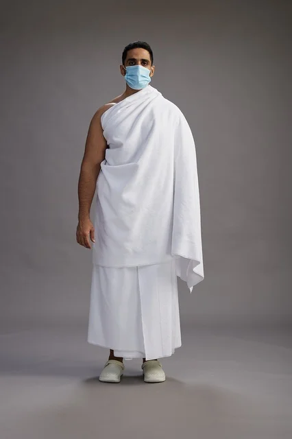 An Arab Saudi Gulf man in Ihram clothing wearing a mask standing ready for Hajj, protection from the coronavirus, full body, gray background.