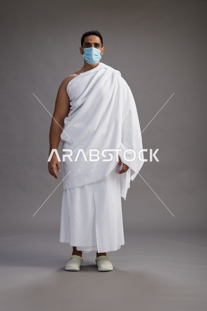 رجل عربي  سعودي  خليجي بملابس الإحرام يرتدي كمامة واقف استعدادًا للحج, الوقايه من فايروس كورونا, كامل الجسم, خلفيه رماديه