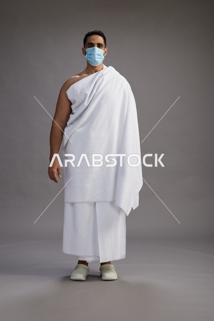 رجل عربي  سعودي  خليجي بملابس الإحرام يرتدي كمامة واقف استعدادًا للحج, الوقايه من فايروس كورونا, كامل الجسم, خلفيه رماديه