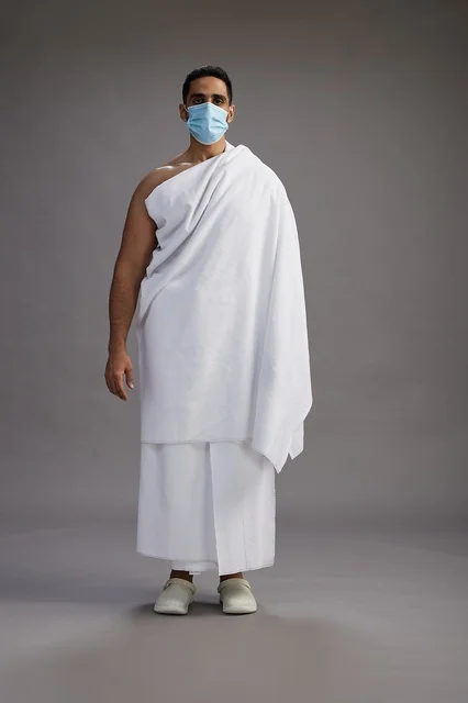 رجل عربي  سعودي  خليجي بملابس الإحرام يرتدي كمامة واقف استعدادًا للحج, الوقايه من فايروس كورونا, كامل الجسم, خلفيه رماديه