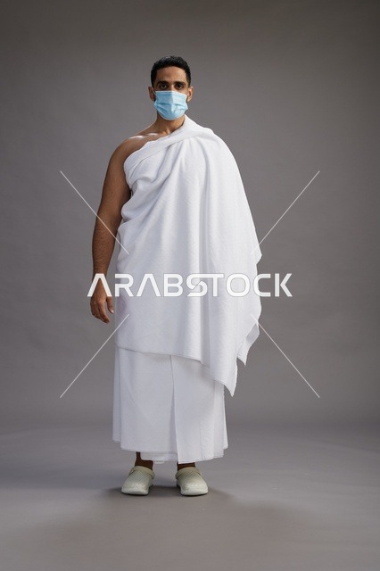 رجل عربي  سعودي  خليجي بملابس الإحرام يرتدي كمامة واقف استعدادًا للحج, الوقايه من فايروس كورونا, كامل الجسم, خلفيه رماديه