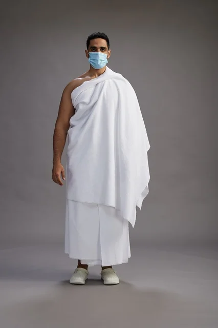 رجل عربي  سعودي  خليجي بملابس الإحرام يرتدي كمامة واقف استعدادًا للحج, الوقايه من فايروس كورونا, كامل الجسم, خلفيه رماديه