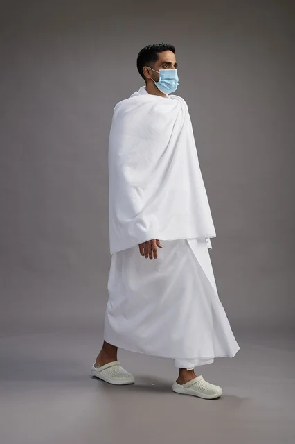 رجل عربي  سعودي  خليجي بملابس الإحرام يرتدي كمامة واقف استعدادًا للحج, الوقايه من فايروس كورونا, كامل الجسم, خلفيه رماديه