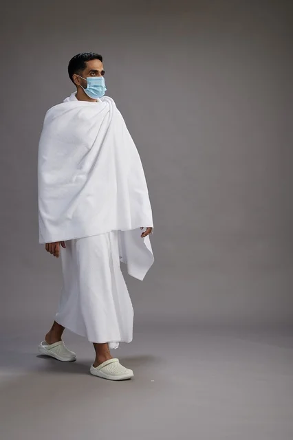 An Arab Saudi Gulf man in Ihram clothing wearing a mask standing ready for Hajj, protection from the coronavirus, full body, gray background.