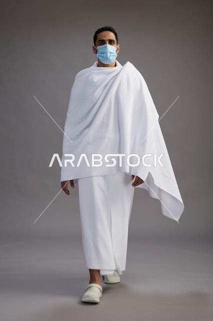 رجل عربي  سعودي  خليجي بملابس الإحرام يرتدي كمامة واقف استعدادًا للحج, الوقايه من فايروس كورونا, كامل الجسم, خلفيه رماديه