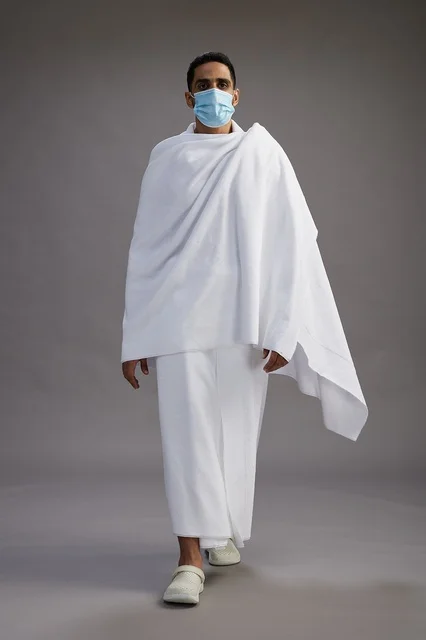 رجل عربي  سعودي  خليجي بملابس الإحرام يرتدي كمامة واقف استعدادًا للحج, الوقايه من فايروس كورونا, كامل الجسم, خلفيه رماديه