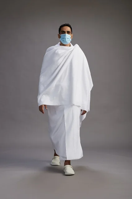 An Arab Saudi Gulf man in Ihram clothing wearing a mask standing ready for Hajj, protection from the coronavirus, full body, gray background.
